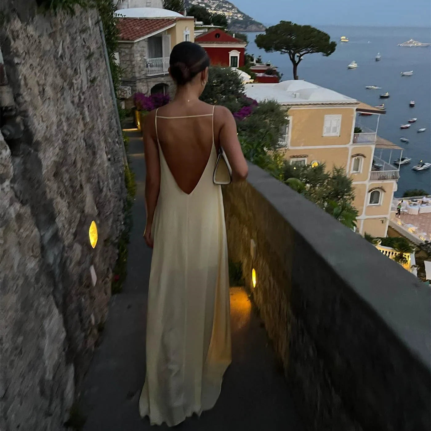 Woman in elegant backless dress walking along scenic coastal pathway with sea view and villas