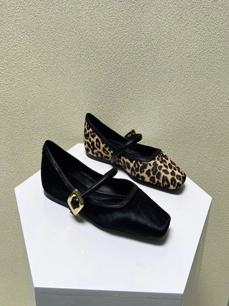 Concierge Tiers displayed with two flat Mary Jane shoes, one black with gold buckle and one leopard print, on a white pedestal