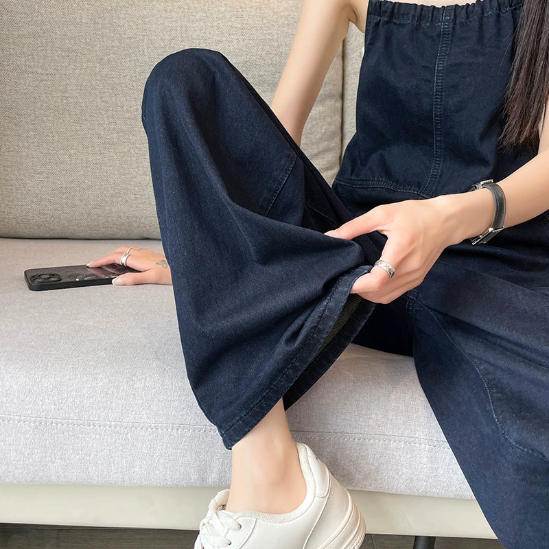 Woman in loose dark denim jumpsuit and white sneakers sitting on a sofa, holding a phone